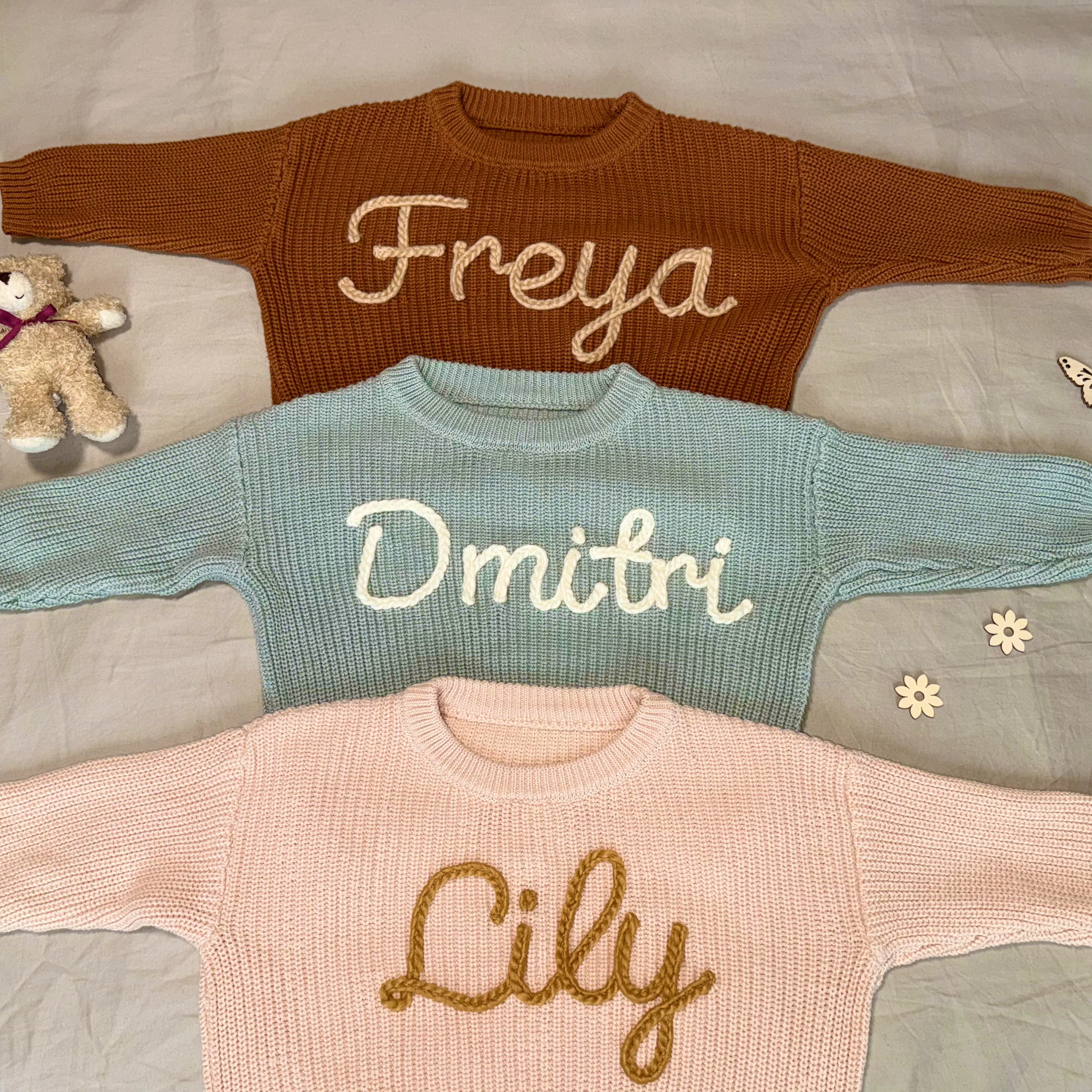 Three knitted sweaters with embroidered names on a bed