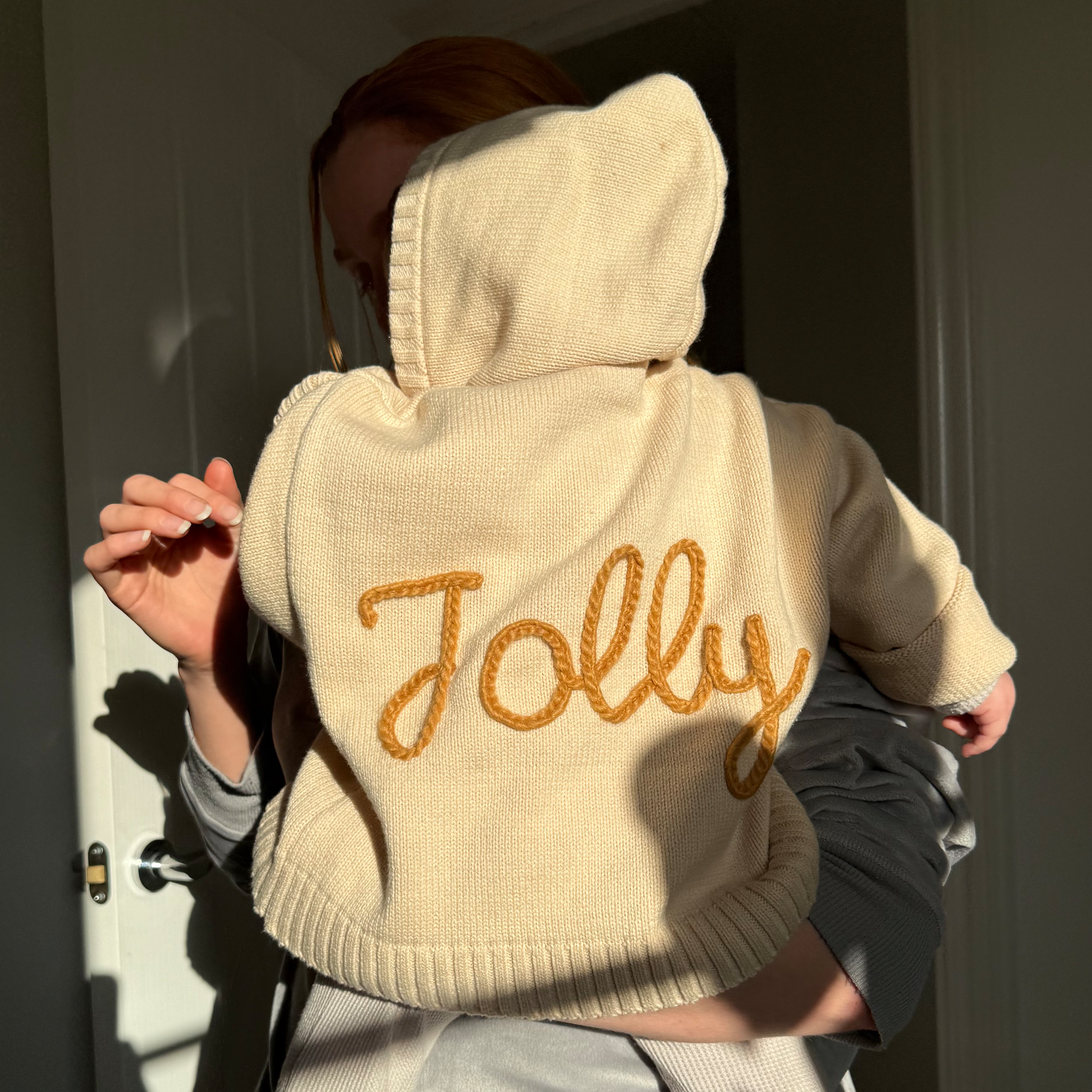 Baby wearing a beige hoodie with 'Jolly' embroidered on the back, standing in a doorway.